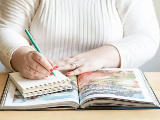 Frau schreibt mit grünem Stift in Notizbuch, während sie ein aufgeschlagenes Fotoalbum betrachtet. Sie erstellt ihr persönliches Jahrbuch.