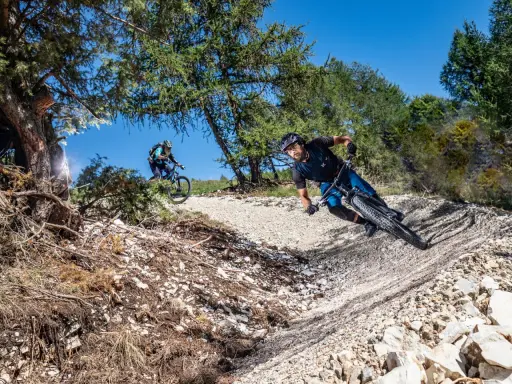 Due ciclisti affrontano un sentiero sterrato in discesa, immersi nella natura. La **tonezza del Cimone** si percepisce nell'aria fresca e nel paesaggio montano.
