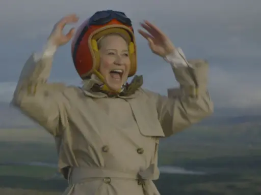 Glad Trine Dyrholm i pilotmössa och trenchcoat vinkar glatt. En scen från filmer med Trine Dyrholm.