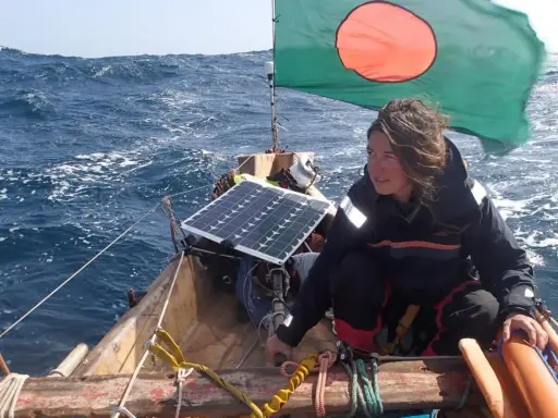 Capucine Trochet navigue sur une mer agitée, le drapeau du Bangladesh flottant au vent. Un panneau solaire est visible sur le bateau.