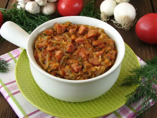 Pyszny bigos z młodej kapusty z kiełbasą, podany w białej misce na zielonym talerzyku. W tle pomidory, czosnek i koperek.