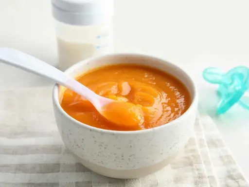 Un tazón con puré de calabaza bebé, una cuchara rosa y un biberón.