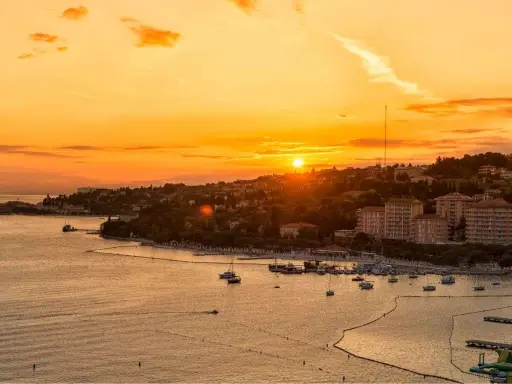 Tramonto dorato sul mare in Slovenia, con un porto pieno di barche e una costa verdeggiante. Immagina i bambini che giocano felici sulla spiaggia.