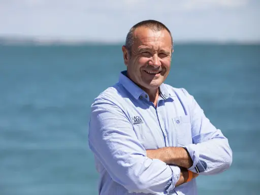 Lalou Roucayrol, souriant, bras croisés, devant la mer. Il porte une chemise bleue rayée avec un logo sur la poitrine.