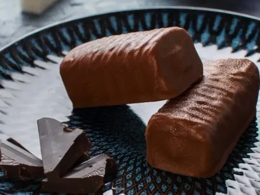 Dwa czekoladowe batony proteinowe na talerzu, obok kawałki gorzkiej czekolady. Idealne do domowego batony proteinowe przepis.