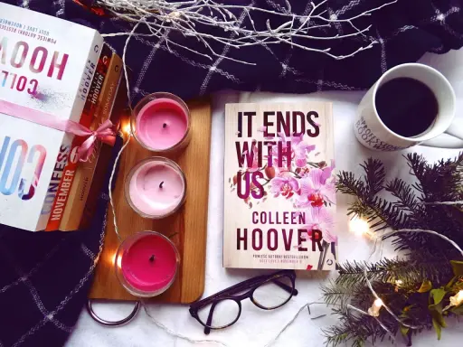 Książka "It Ends With Us" Colleen Hoover, obok świec, kawy i innych książek, tworzy przytulną atmosferę do czytania.
