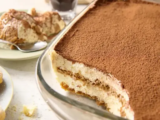 Pyszne tiramisu bez jajek w szklanej formie, posypane kakao. Obok porcja deseru na talerzyku.