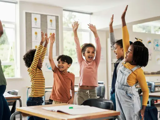 Fröhliche Kinder mit erhobenen Armen in einem Klassenzimmer. Sie nehmen begeistert an Kennenlernspielen für die Grundschule teil.