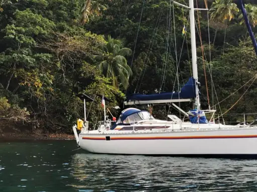 Un voilier blanc, un voilier Jurançon, est ancré dans une baie tropicale luxuriante, entouré d'une végétation dense et de palmiers.