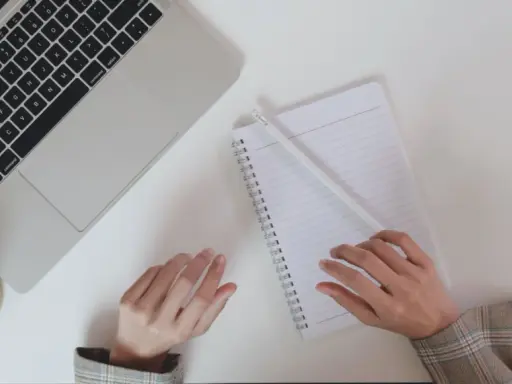 Ręce piszą notatki do CV po niemiecku przy laptopie, obok filiżanka kawy.