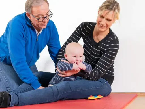Ein Mann und eine Frau üben mit einem Baby auf einer roten Matte. Das Baby greift nach seinen Fingern. kg zns kinder.