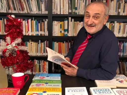 Daniele Novara, autore di libri per genitori, legge un suo libro tra scaffali pieni di volumi, con un albero di Natale e una bottiglia di spumante.