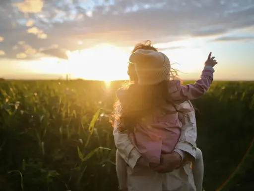 Bambina aggrappata alla schiena di un adulto, con il sole al tramonto che illumina un campo. Un'immagine di puro attaccamento affettivo.