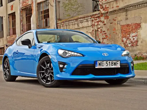 Błękitna Toyota GT86, znana też jako Toyota BRZ, zaparkowana przed zniszczonym budynkiem.