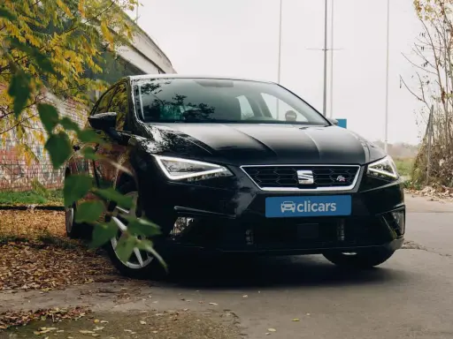 Coche negro SEAT León FR aparcado entre hojas secas y un muro de ladrillos. El emblema "FR" indica su versión deportiva.
