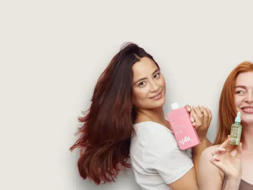 Kobiety prezentują kosmetyki do włosów. Jedna trzyma różową butelkę, druga małą buteleczkę z serum, które zawiera olejek jojoba właściwości odżywcze.
