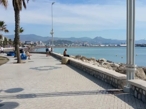 Ein Mann sitzt am Ufer der playa de la misericordia und blickt auf das Meer. Palmen säumen die Promenade, im Hintergrund die Stadt und Berge.