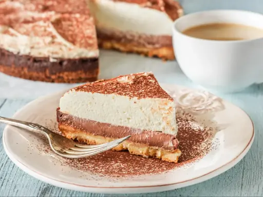 Pyszna tarta z mascarpone i czekoladą, posypana kakao, czeka na widelczyk. Obok filiżanka kawy.