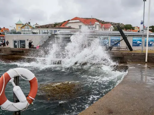 Vågor slår mot kajen i en kuststad, med hus i bakgrunden. En livboj syns i förgrunden. Det blåser med sekundmeter storm.