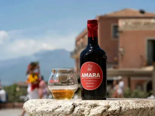 Bottiglia e bicchiere di Amara, un amaro che celebra come l'arancio amaro trama i sapori siciliani. Sfondo sfocato di un borgo.