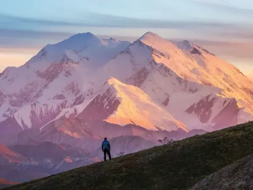 Widok na najwyższy szczyt Ameryki Północnej, Denali, oświetlony zachodzącym słońcem. Samotny turysta stoi na zboczu, podziwiając majestat góry.