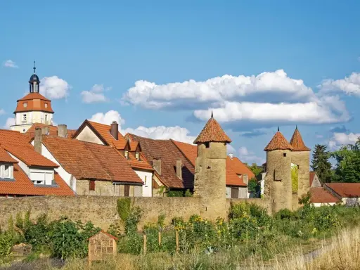 Eine malerische **Stadt im Mittelalter** mit roten Dächern, steinernen Türmen und einer alten Stadtmauer unter blauem Himmel.