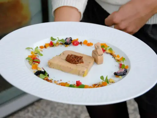 Une présentation raffinée de foie gras en entrée, ornée de fruits secs, de baies et de pousses vertes sur une assiette blanche texturée.