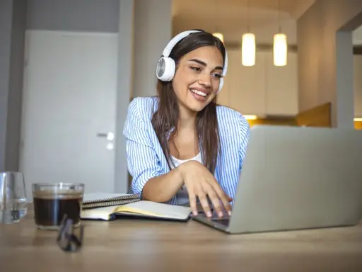 Uśmiechnięta studentka w słuchawkach pracuje przy laptopie. Czy muzyka pomaga w nauce? Ona chyba tak, bo wygląda na zrelaksowaną i skupioną.