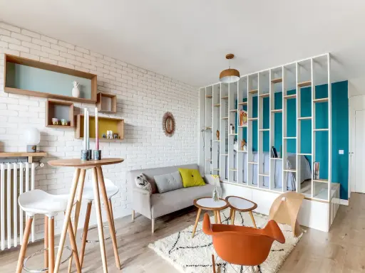 Idée aménagement studio 25m2 : un espace salon cosy avec canapé, table haute et étagères murales, séparé d'une chambre par une bibliothèque ajourée.