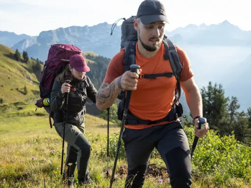 Dos excursionistas con mochilas y bastones caminan por una ladera montañosa.