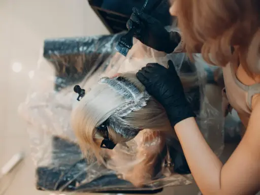Fryzjerka w czarnych rękawiczkach nakłada farbę na blond włosy. Kluczowe jest, ile trzymać farbę na włosach, by uzyskać idealny efekt.