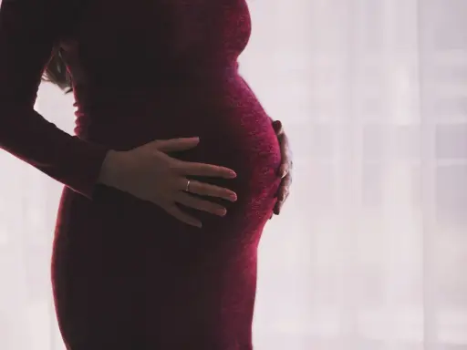 Kobieta w ciąży w bordowej sukience delikatnie obejmuje dłońmi brzuszek.