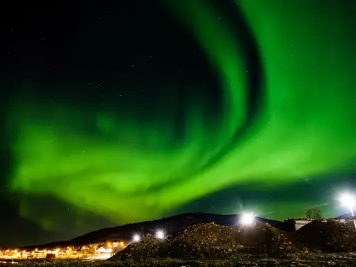 Spektakularna zorza polarna rozświetla nocne niebo nad ośnieżonymi wzgórzami i światłami miasteczka. To idealny moment, kiedy występuje zorza polarna.
