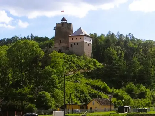 Zamek w Czchowie góruje nad zielonym wzgórzem, otoczony drzewami. Na szczycie wieży powiewa flaga.