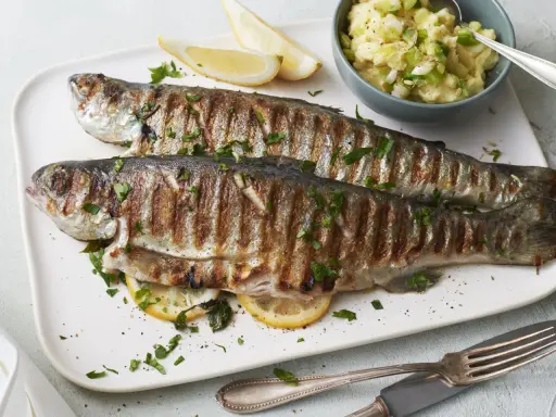 Zwei perfekt gegrillte Forellen liegen auf einem Teller, garniert mit Petersilie und Zitronenspalten. Dazu gibt es Kartoffelpüree mit Frühlingszwiebeln. Ein Genuss für alle, die Forelle grillen lieben.