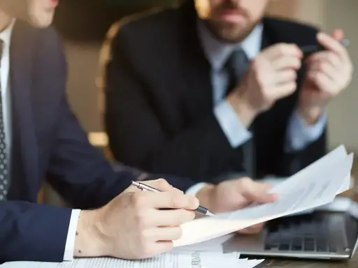 Dwóch mężczyzn w garniturach analizuje dokumenty. Jeden z nich wskazuje piórem na umowę, która może być naganą dla pracownika.