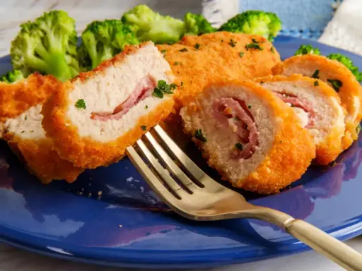 Kotlet po szwajcarsku, panierowany, z szynką w środku, podany z brokułami na niebieskim talerzu. Złoty widelec czeka na pierwszy kęs.
