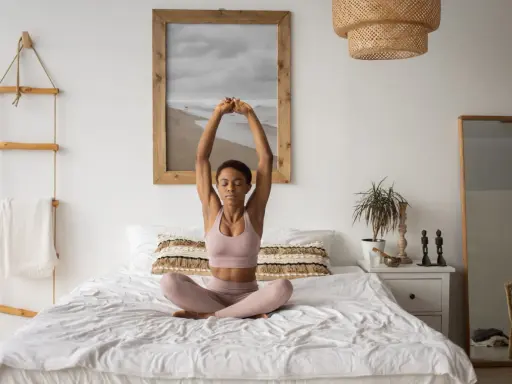 Frau praktiziert Yoga im Bett, mit geschlossenen Augen und erhobenen Armen. Ein Bild von einem Strand hängt an der Wand.