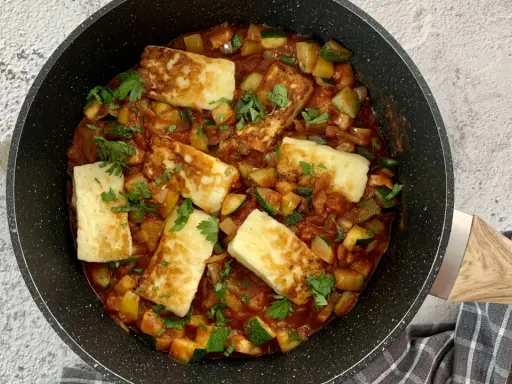 Smażony ser halloumi z warzywami w sosie pomidorowym, posypany natką pietruszki, w czarnej patelni.