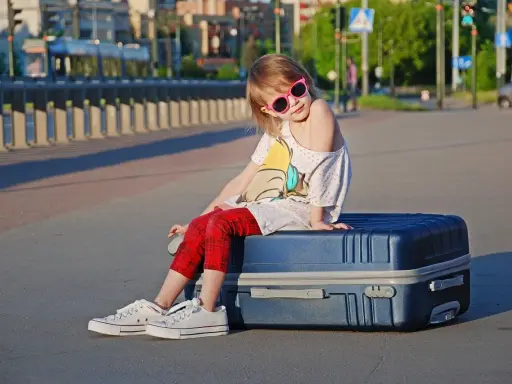Dziewczynka w okularach siedzi na walizce, gotowa na wyjazd dziecka za granicę.