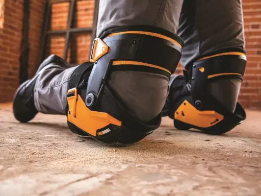 Protection des genoux pour éviter toute maladie professionnelle. Des genouillères robustes et orange protègent les genoux d'un travailleur sur un sol poussiéreux.