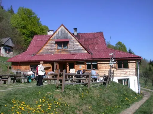 Schronisko pod Bereśnikiem, drewniany budynek z czerwoną dachem, otoczony zielenią. Kilka osób odpoczywa na ławkach przed schroniskiem.