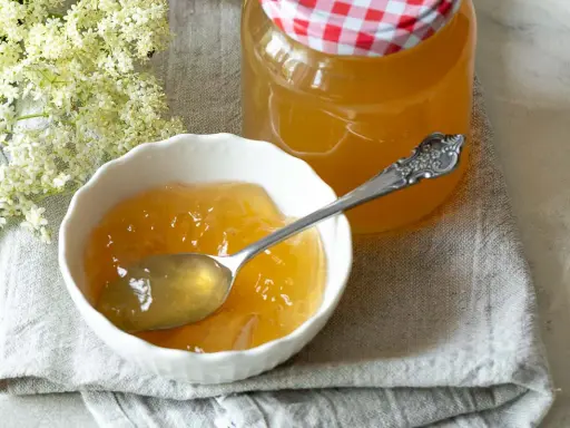 Ein Löffel voll goldgelbem Holunder Apfel Gelee in einer Schale, daneben ein Glas mit dem Gelee und Holunderblüten.