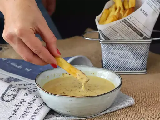 Une frite trempée dans une sauce moutarde miel onctueuse. Des frites dorées dans un panier en métal à côté.