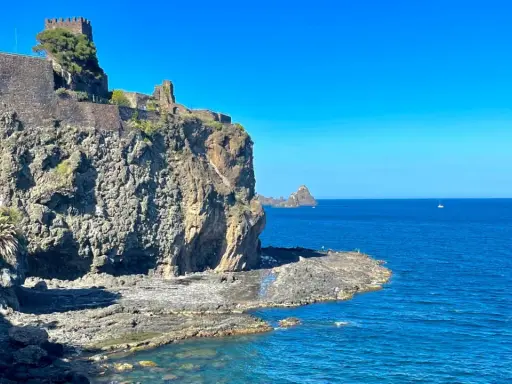 Castello Normanno a Aci Trezza, un luogo da vedere. Roccioso promontorio sul mare blu, con un antico castello in pietra e vegetazione.