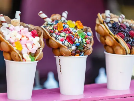 Trois bubble waffles colorés, garnis de bonbons, guimauves et fruits, présentés dans des gobelets blancs.