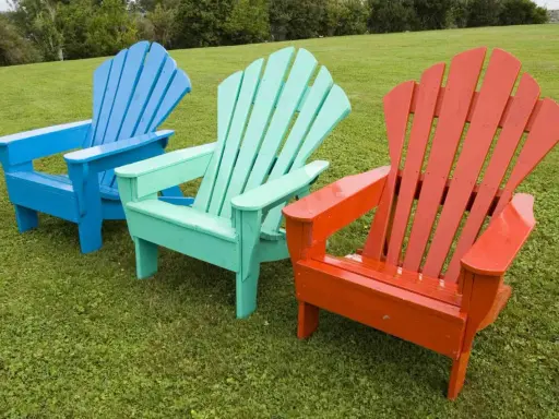 Tres sillas de madera de estilo Adirondack, pintadas de azul, verde menta y naranja, lucen vibrantes sobre el césped. ¡Un proyecto de pintar sillas de madera sin lijar que da vida al jardín!