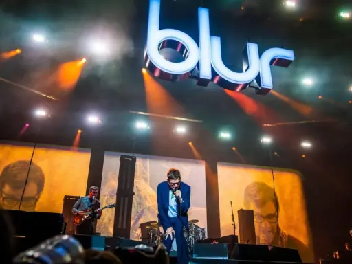 Medlemmar av Blur uppträder på scenen under en konsert, med den ikoniska logotypen lysande ovanför.
