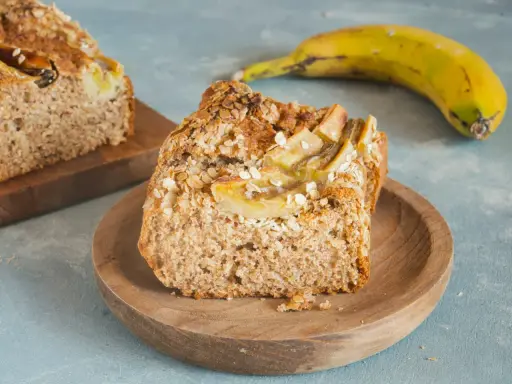 Pyszny chlebek bananowy fit z płatkami owsianymi na wierzchu, podany na drewnianym talerzyku. Obok leży dojrzały banan.