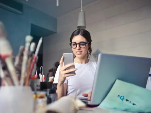 Kobieta w okularach do komputera przegląda telefon, siedząc przy laptopie.